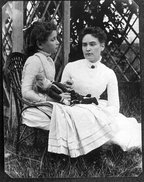 A vintage photo shows a young girl, Helen Keller, seated in a chair while her adult teacher, Anne Sullivan, reclines on the ground next to her. Both women are dressed in white. Helen holds a doll in her lap.