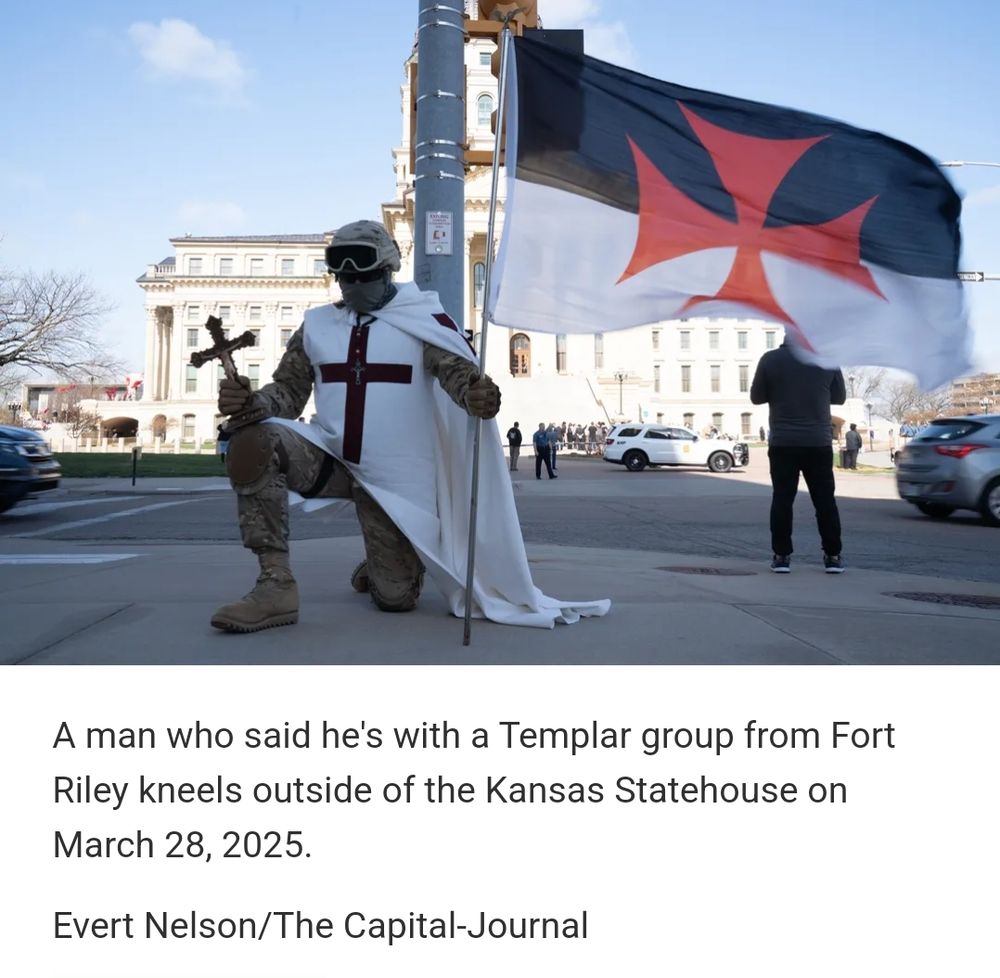 A man dressed in military camo and boots wears sun glasses, a mask, and a helmet with goggles pulled up. Above this he wears a white tunic and robe with a large, brown cross on the front. He is taking a knee across the street from the Statehouse. In his right hand he grasps a large cross, in his right hand he holds a bicolored flag. The top half is dark blue and the bottom half is white. Stitches upon the flag is a red x-like symbol reminiscant of the old crusades. A police car and police stand near a group of protesters on the capitol driveway. 