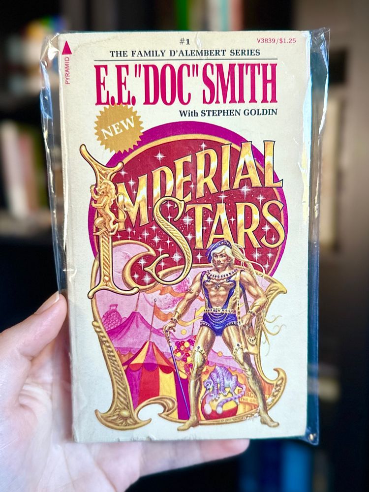 The cover of E.E. “Doc” Smith’s “Imperial Stars”, which features the most golden, glittering, homosexual lettering you ever did see, over a bunch of pink circus tents.  Before the tents stands a shirtless, shiny blonde man in what looks like tight purple-and-gold bondage wear and thigh-high gold boots.  He’s got an absolutely swagtastic mustache.