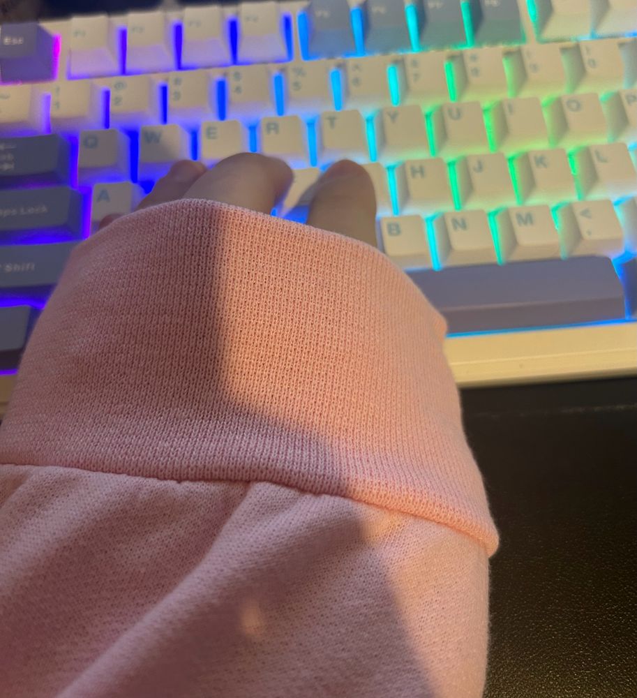 The image shows a close-up of my hand resting on a brightly lit mechanical keyboard. The hand is partially covered by the sleeve of a soft pink sweatshirt.