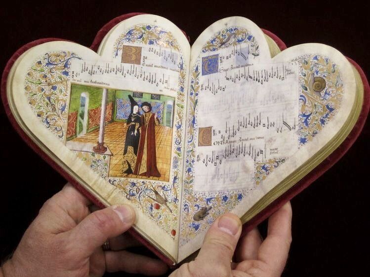 The 15th century chansonnier of Jean de Montchenu, an illuminated manuscript with heart-shaped pages and binding. A person's hands hold it open to reveal musical notations and an illustration of a man and woman walking together.