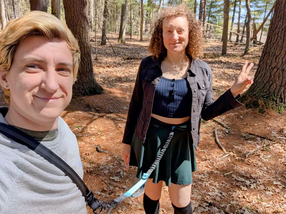 Mylo takes a selfie of themselves leading Violet Rollergirl through a casual walk in some woods. A harness leash is around their waist and attached to Violet's waist.