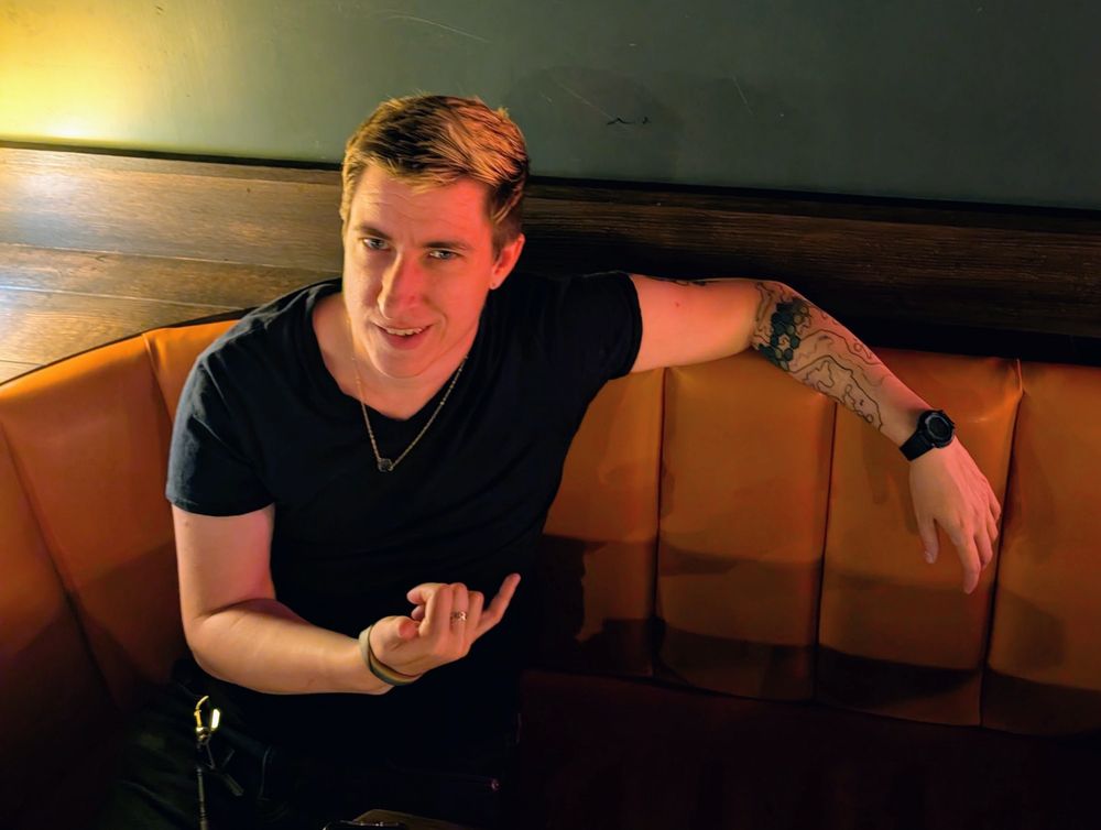 Mylo Ryn gives a shaka hand 🤙🏻 and flashes their confident and charming smile at you while sitting in an empty booth at a darkly lit venue.