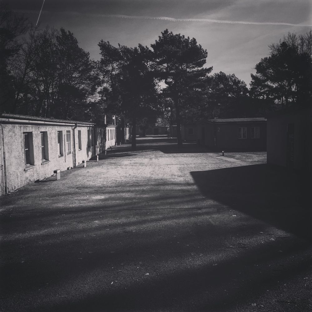 Foto der Gedenkstätte NS-Zwangsarbeiterlager Berlin Schöneweide, Februar 2019. Schwarz-Weiß-Aufnahme des Innenhofs zwischen den Baracken mit langen Schlagschatten, am Himmel zwei sich kreuzende Kondensstreifen