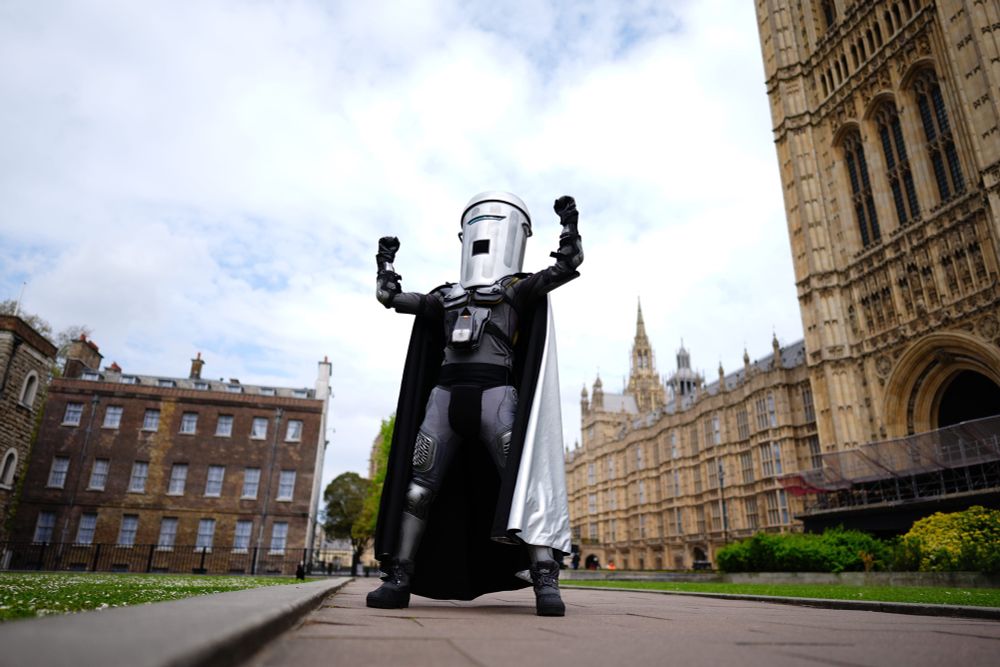 Count Binface, a costumed man in black wearing a silver floor-length cape and a garbage can on his head stands in front of the British parliament building with his feet wide and fists triumphantly in the air
