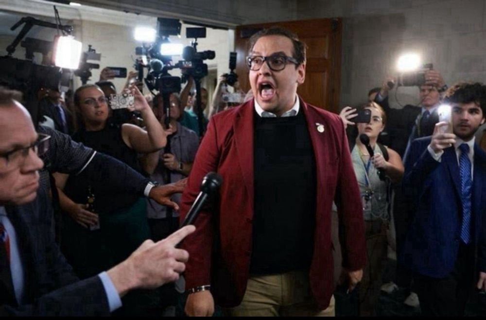 George Santos upon being expelled from congress. He is surrounded by reporters with microphones and cameras, and his mouth is dramatically open so it looks like he is shouting or singing.