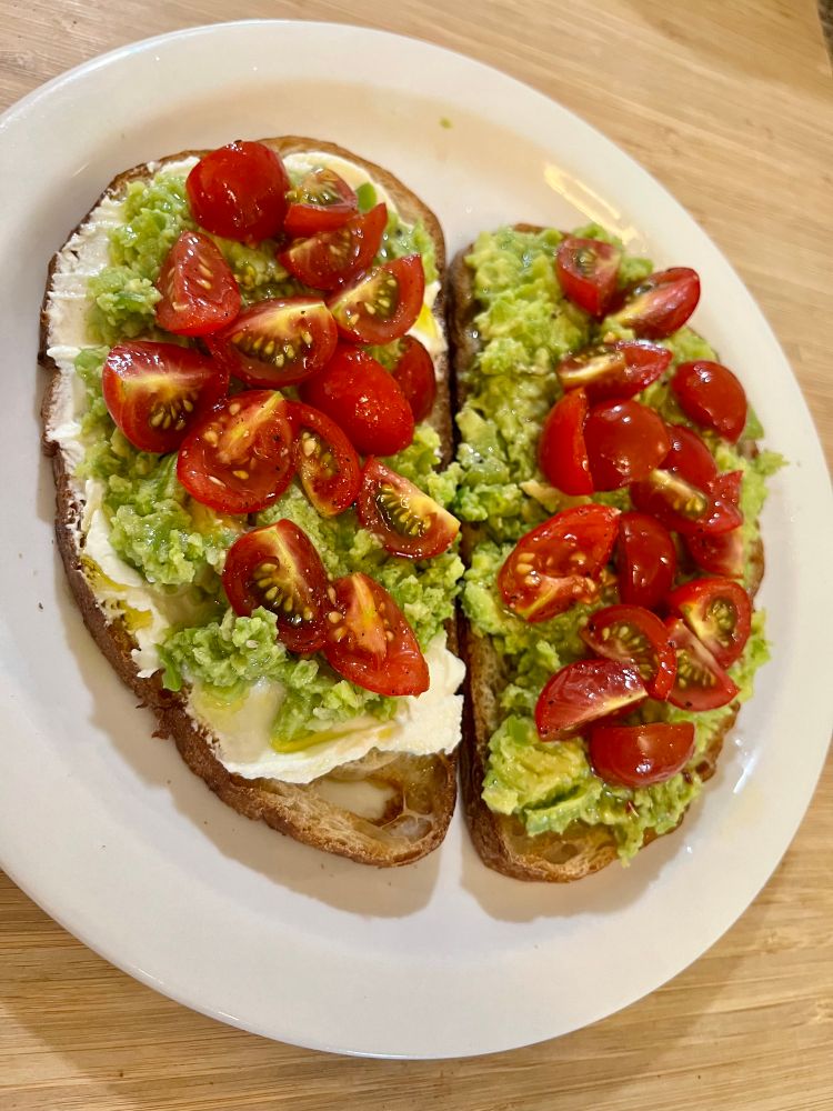 Two slices of toasted bread topped with ricotta, fava bean avocado mash, and cherry tomato wedges on a white plate. 