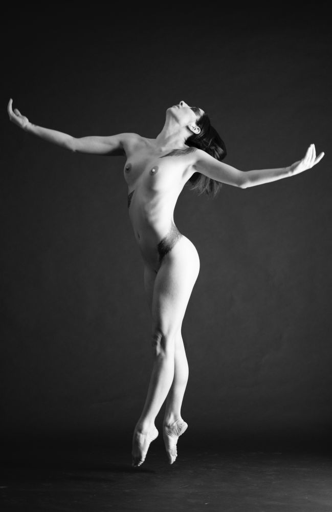 A dancer, nude with a black studio background. She is springing from the floor en pointe, arms wide, chest proud, palms up, chin raised. Lit from the right, body angled slightly away from the light. 