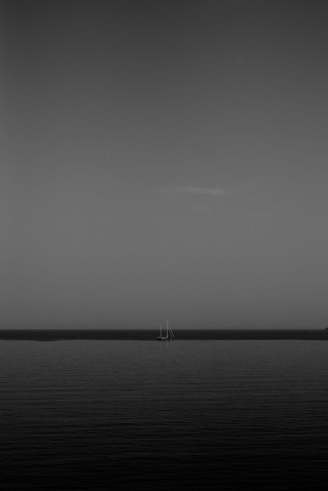 A calm sea, expansive, a clear sky cloudless aside from one visible whisp. Just before the horizon, centred in the image, a sailing boat, sails down. 