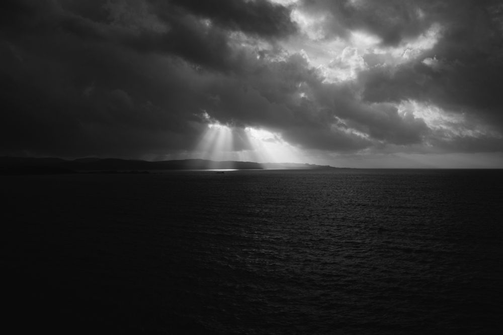 A black and white photograph of the ocean. Land in sight, far away, heavy cloud cover. 2 sections in the clouds where beams of the suns rays have broken through and are illuminating the water below. 