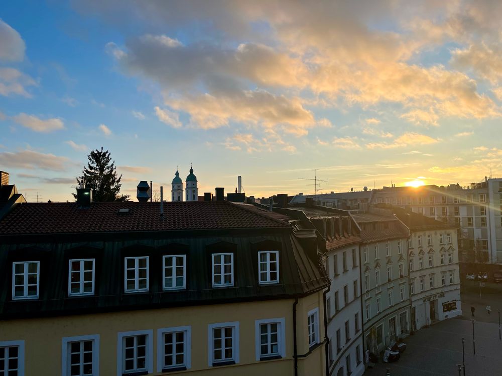 Blick aus dem Fenster auf einen Platz, Bürgerhäuser im Halbrund, Abendlicht, im Hintergrund zwei Kirchtürme und eine Tanne.