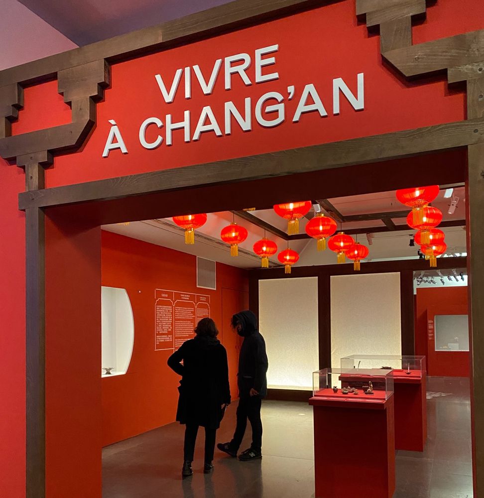 Entrée dans une salle d’exposition. Au premier plan, un portail rouge avec écrit « Vivre à Chang’an » sur le frontispice. A l’arrière plan, une salle où sont exposés des artefacts, et où se trouvent deux visiteurs en train de lire un texte explicatif écrit sur un mur. Au plafond, des lanternes Chinoises servent de décoration.