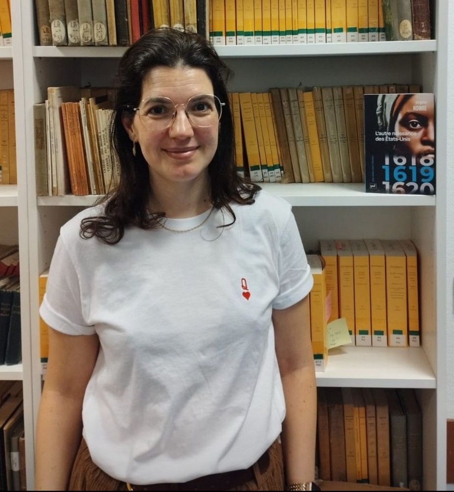Portrait de Virginie Adane devant une une bibliothèque blanche où se trouvent des livres scolaires. Sur un des étages du haut à droite est posé le livre 1619.