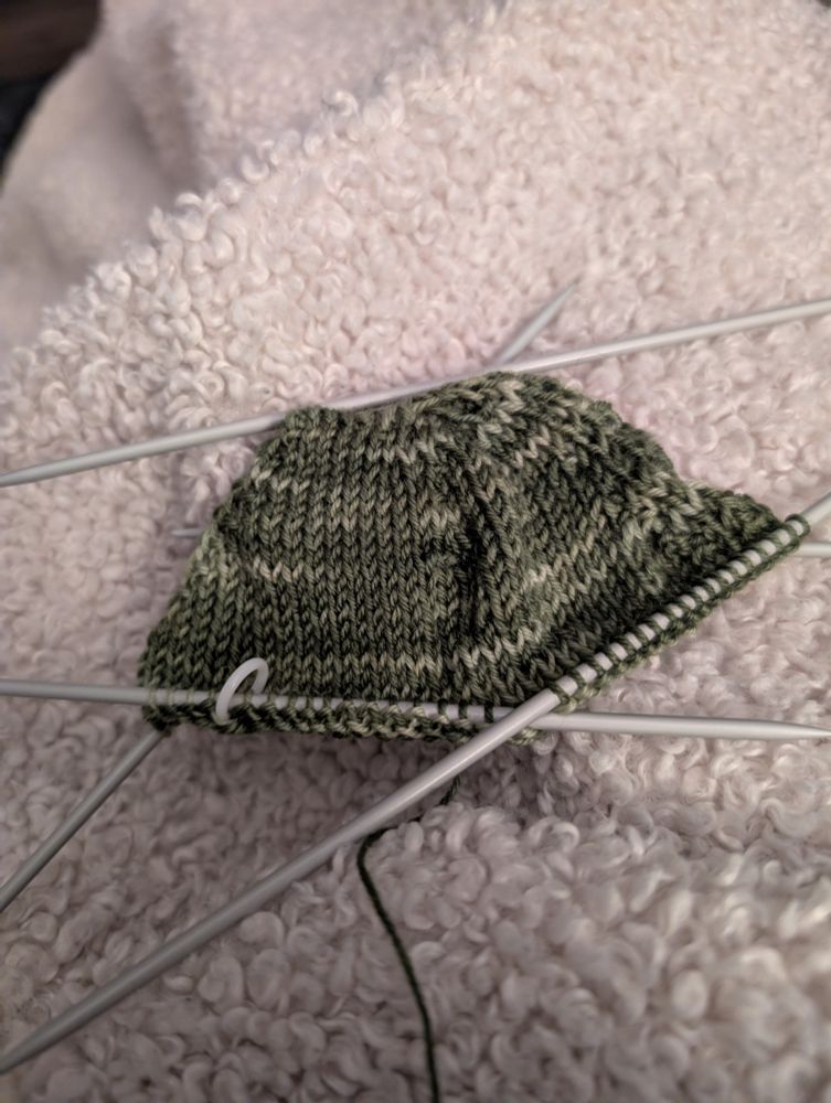 A small bit of green wool knitted into the toe of a sock. It is on four double pointed needles, with a fifth resting to the side. 