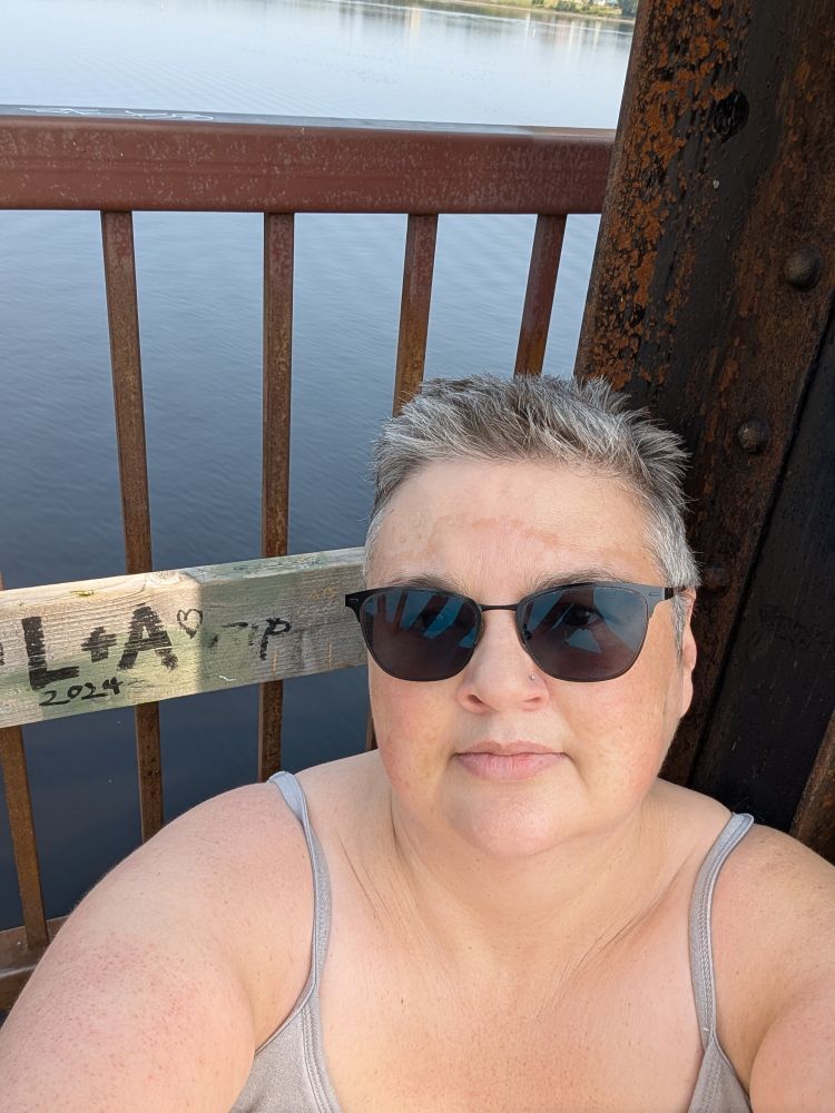Julie, a white person with short brown and grey hair wearing sunglasses and a taupe spaghetti strap top, sits at the side of a rusted rail bridge over the Wolastoq River. 