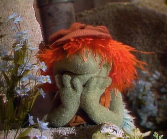 Boober Fraggle, a green puppet with bright orange hair wearing an orange cap, stands leaning on a rock with his chin in his hands. 