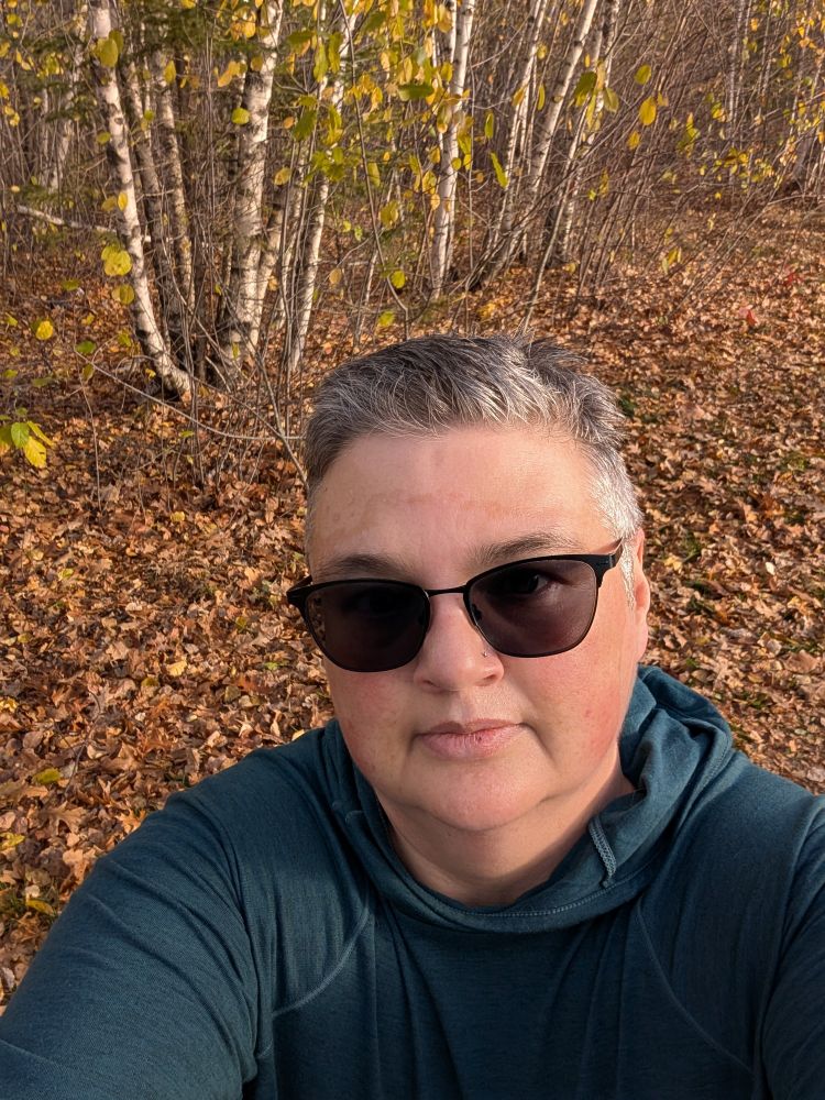 Julie, a white person with short brown and grey hair wearing sunglasses and a teal hoodie, stands in front of a bunch of birch trees. There's brown leaves all over the forest floor. 