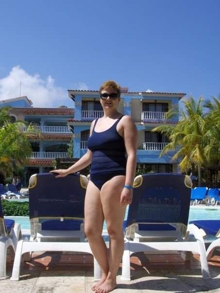 Julie, a white person with short brownish red hair wearing a navy one piece swimsuit and sunglasses, stands next to a lounge chair near a resort pool. 