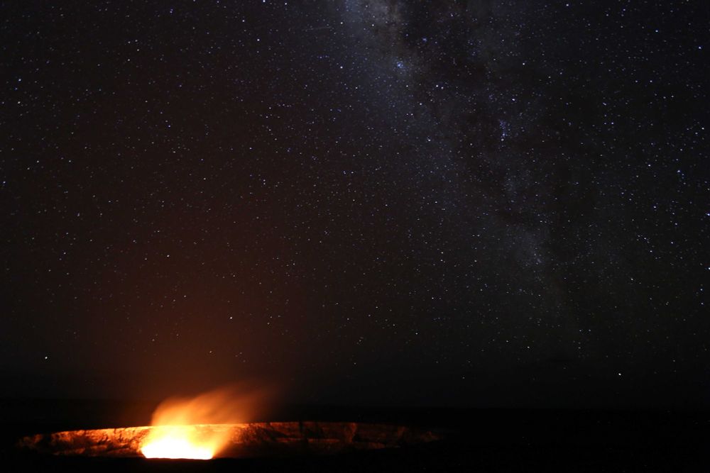 A photograph of the night sky taken at Kīlauea, on Hawaii. The glow of the volcano is visible in the bottom left, lighting up the ridge of the volcanic crater. In the center right of the image, the Milky Way can be seen.