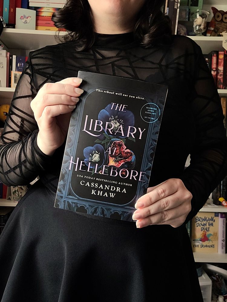 Image of a woman standing in front of bookshelves holding a book titled The Library at Hellebore by Cassandra Khaw. The woman is wearing a black mesh top and a black skirt with their face cropped out of the image. 