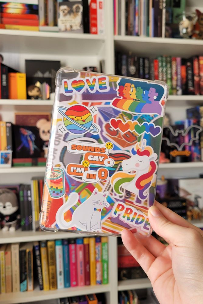 photo of a hand holding up a clear kindle case with a decorative cardstock insert in front of a bookshelf. the decorative insert is pride themed with various different pride stickers placed over a pride background. the stickers are all graphic images made to look real and 3D.