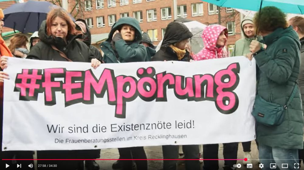 Szene aus der Dokumentation „Macht, Kontrolle, Femizid – Wer schützt Frauen vor Gewalt?“, zu sehen ist eine Gruppe von Demonstrantinnen mit einem Transparent, auf dem steht: #FEMpörung - Wir sind die Existenznöte leid! - Die Frauenberatungsstellen im Kreus Recklinghausen