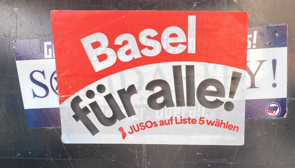 Ein Aufkleber gegen Antisemitismus überklebt von einem Aufkleber JUSO Basel.

Aufkleber der JUSO Basel: Weiss auf rot steht "Basel" darunter schwarz auf weiss "für alle!", kleiner steht darunter in rot auf weiss das Logo der JUSO Schweiz und "JUSOs auf Liste 5 wählen"

Aufkleber gegen Antisemitismus: Ein länglich-breiter Aufkleber, in der Mitte steht gross "SOLIDARITY!" im "O" ist ein Davidsstern. Obendrüber steht "Gegen jeden Antisemitismus!", zuunterst "immer, überall" und ein Antifa-Logo.