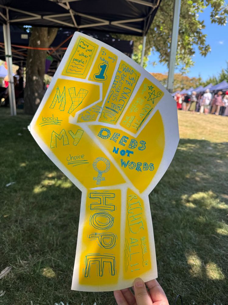 Against the background a field with stalls and people milling about, a large yellow cardboard raised fist placard is decorated with different feminist slogans, designed by my 12 year old daughter. 