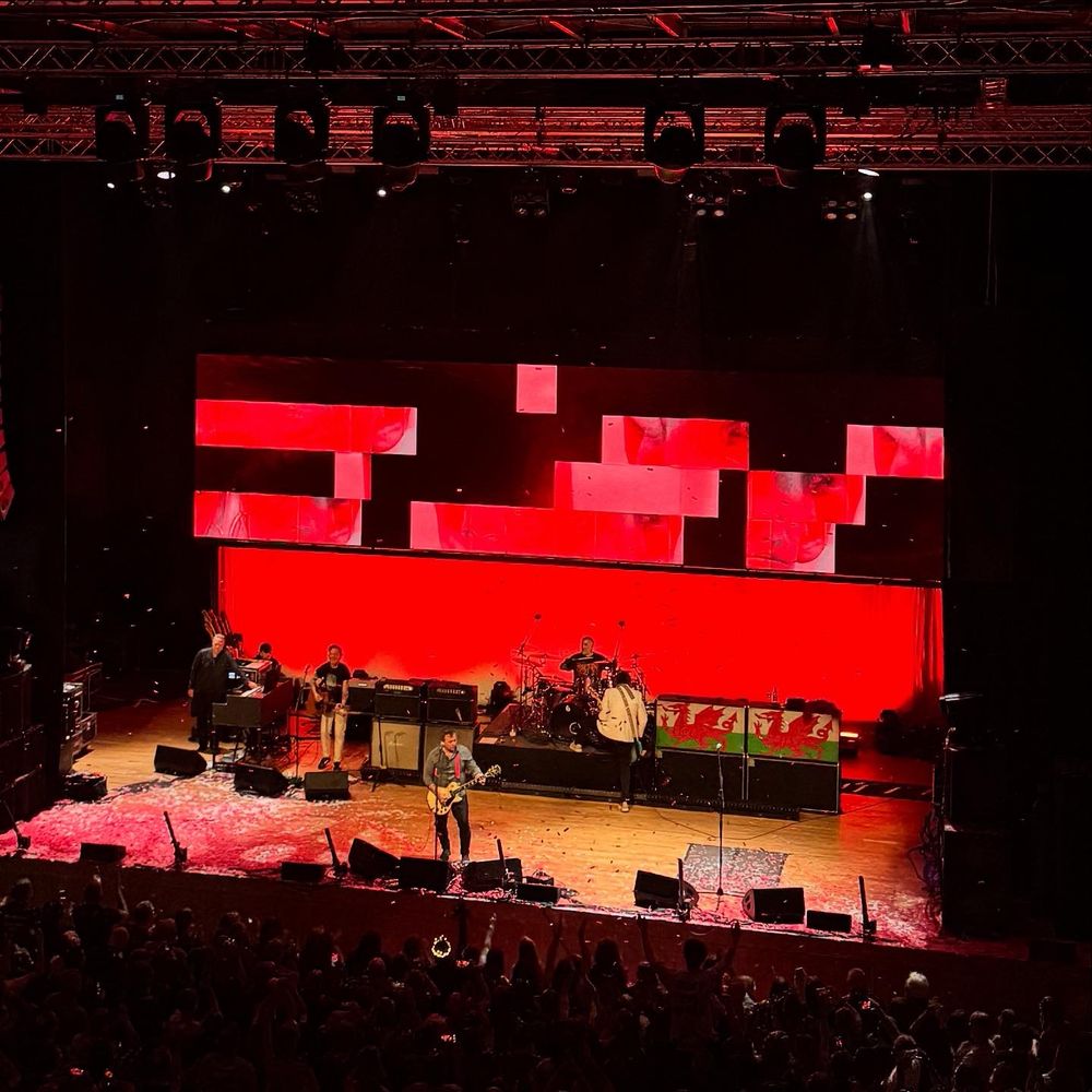 A stage at Wolverhampton civic hall. The lights are down on the crowd and the manic street preachers stand in the stage (three guitarists, drums, keyboard). Red pixelated lights are on the backdrop. Red confetti is on the floor. The lead singer holds a guitar and sings into a microphone. 