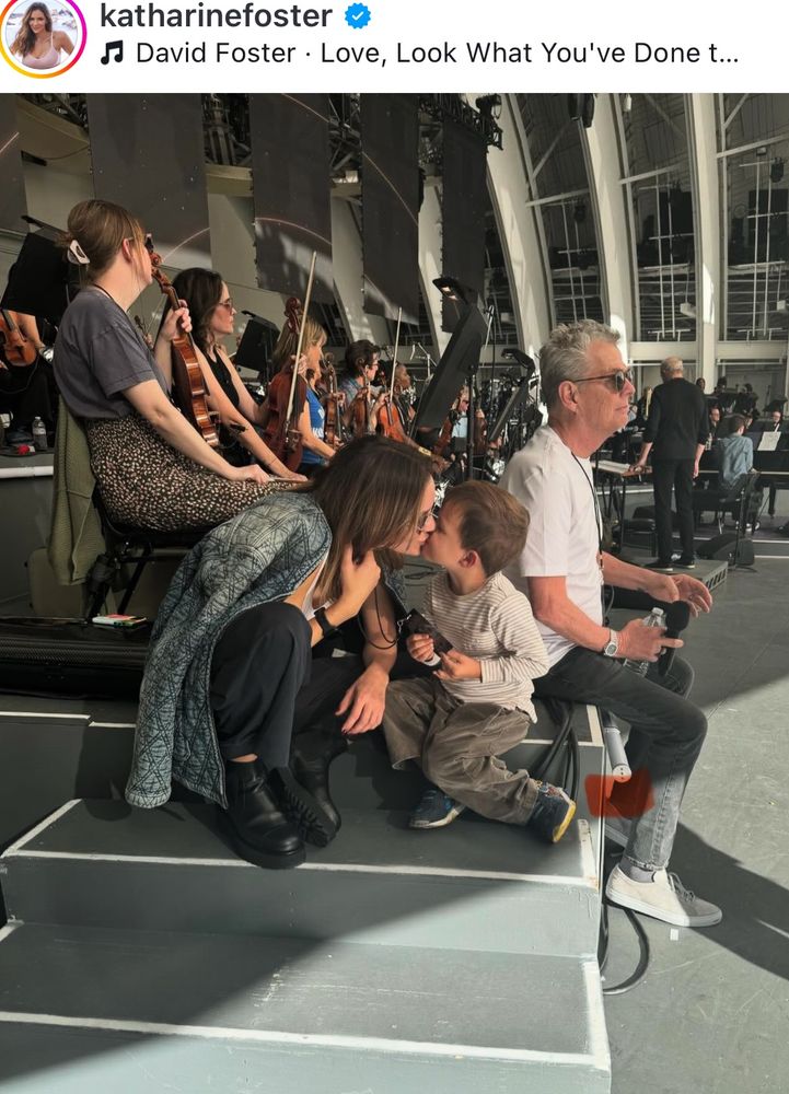 Picture of Katharine Foster bent down kissing her 3yr old son on risers for an orchestra while a side profile of her husband David Foster sits next to them in a white tshirt and faded black jeans and sunglasses. 