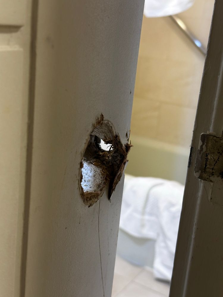 Hole in door where the door handle had been in the door. Bathtub with white towels in the background