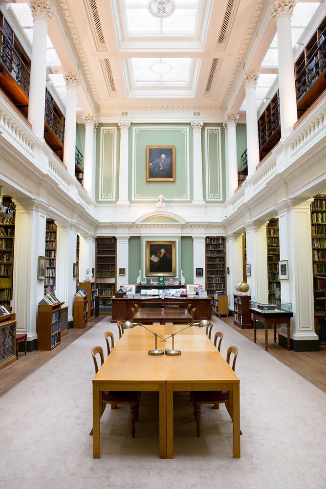 The library of the Linnean Society, with double height pillars and a balcony. The walls are lined with books and a table with lamps is in the centre of the image. There are two paintings, as well as a bust of Linnaeus on the back wall.