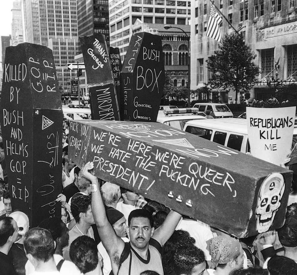 black and white photo of a ACT UP protestors holding up black coffins, main one in foreground “WE'RE HERE, WE'RE QUEER, WE HATE THE FUCKING PRESIDENT” held by visionary writer and poet Assotto Saint (with coffin), protest an appearance by then-president George H.W. Bush, New York City, July 1990. Photo © @dona_ann_mcadams posted by Igbt_history on IG