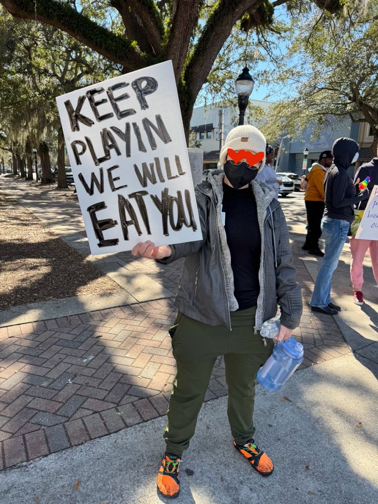 person wearing a mask and eyes blocked out by hearts
holding up a white protest sign with black handwritten lettering
KEEP PLAYIN 
WE WILL EAT YOU
