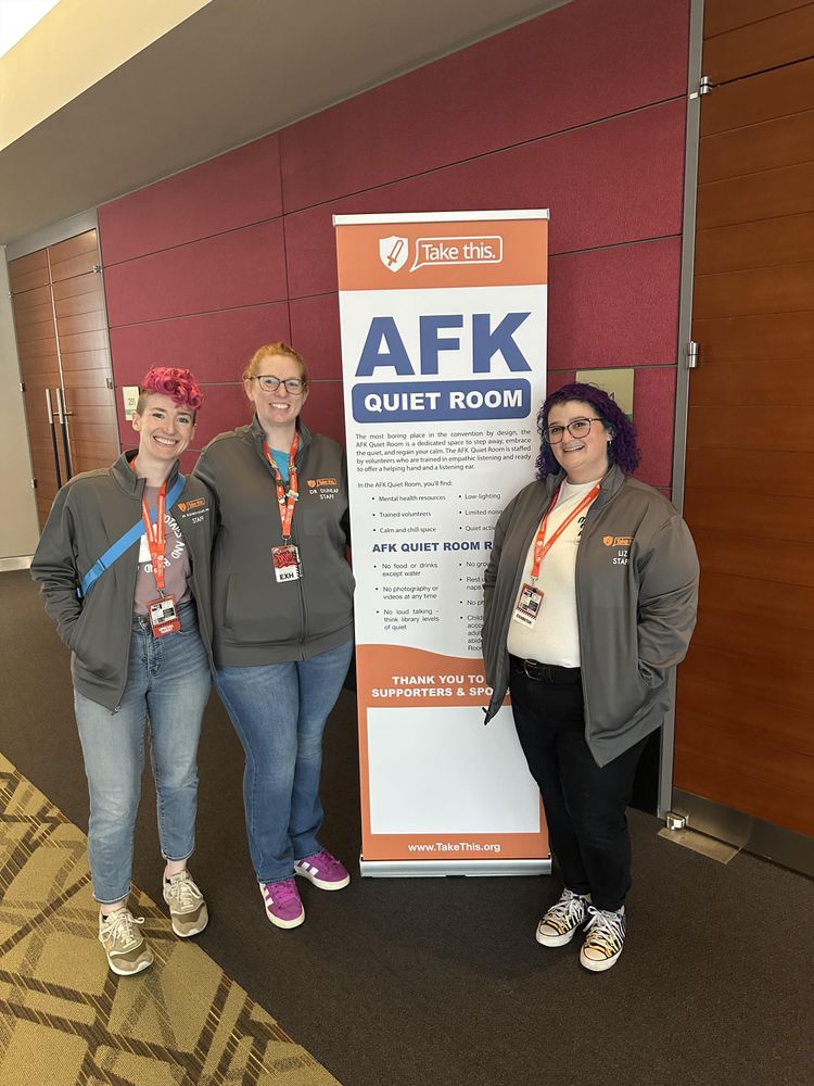 Three Take This staff members, all in grey Take This jackets and wearing convention badges on orange Take This lanyards, are standing in front of an AFK Quiet Room vertical sign with information and rules for the AFK Room. 