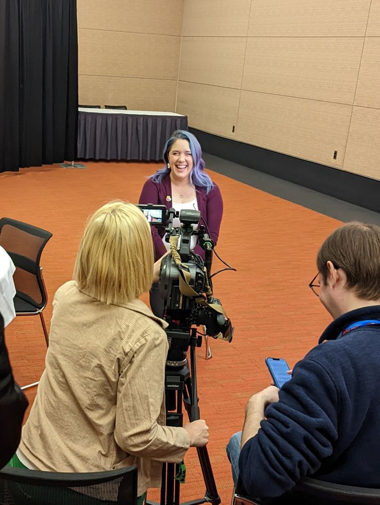 Sarah Hays, a woman with lavender hair wearing a plum sweater with a take this pin on the lapel over a white shirt, sits smiling in front of a camera for an interview. Behind the camera are two individuals with their backs to us, one operating the camera and another looking down at their phone. 
