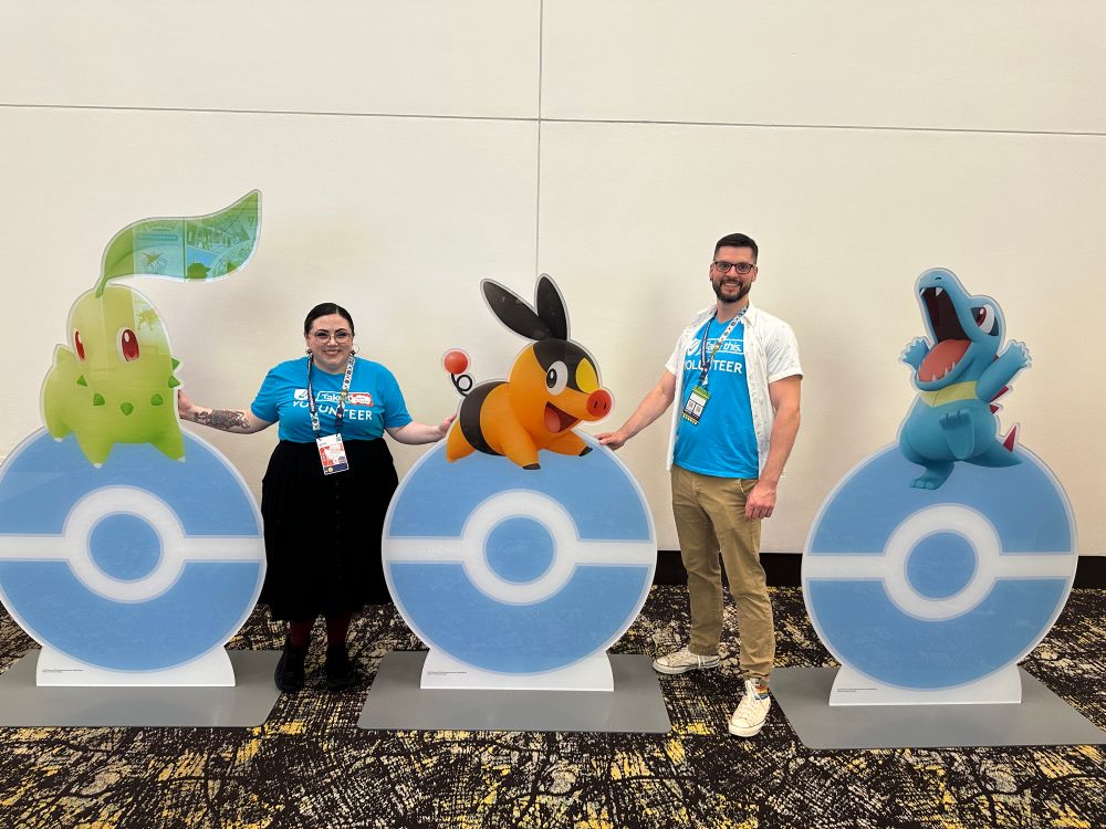 Two take this volunteers stand next to large standees of Chikorita, Tepig, and Totodile