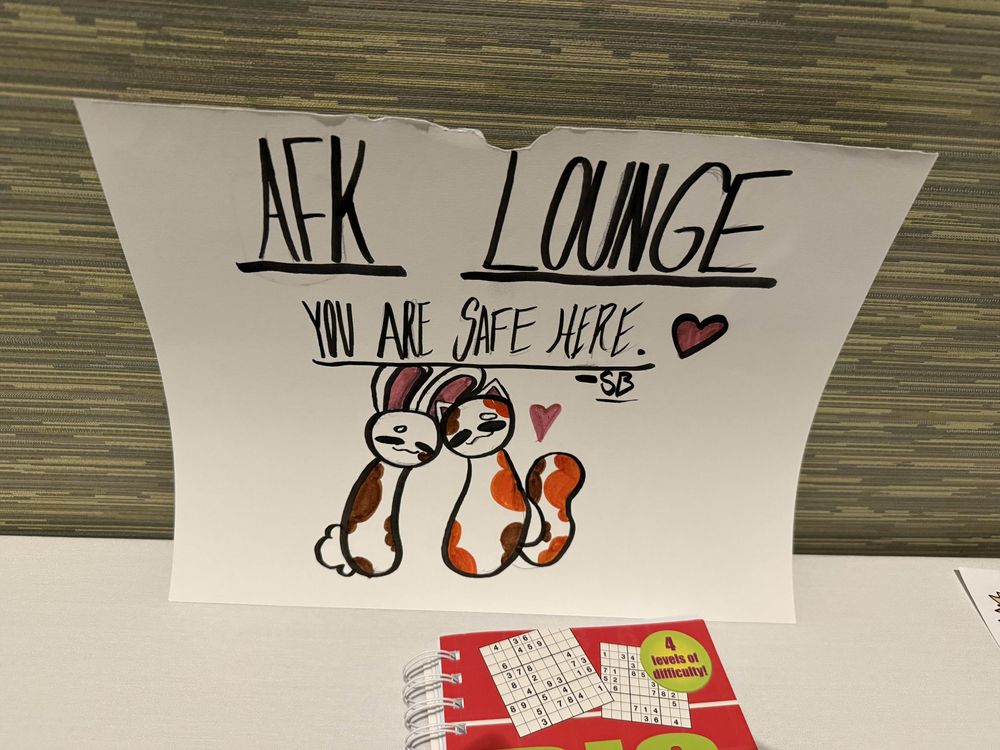 A hand drawn sign. Written text reads "AFK Lounge. You are safe here." Artist signature SB is at the bottom of the text. A rabbit and a cat are below, heads leaned together in a cozy pose. 