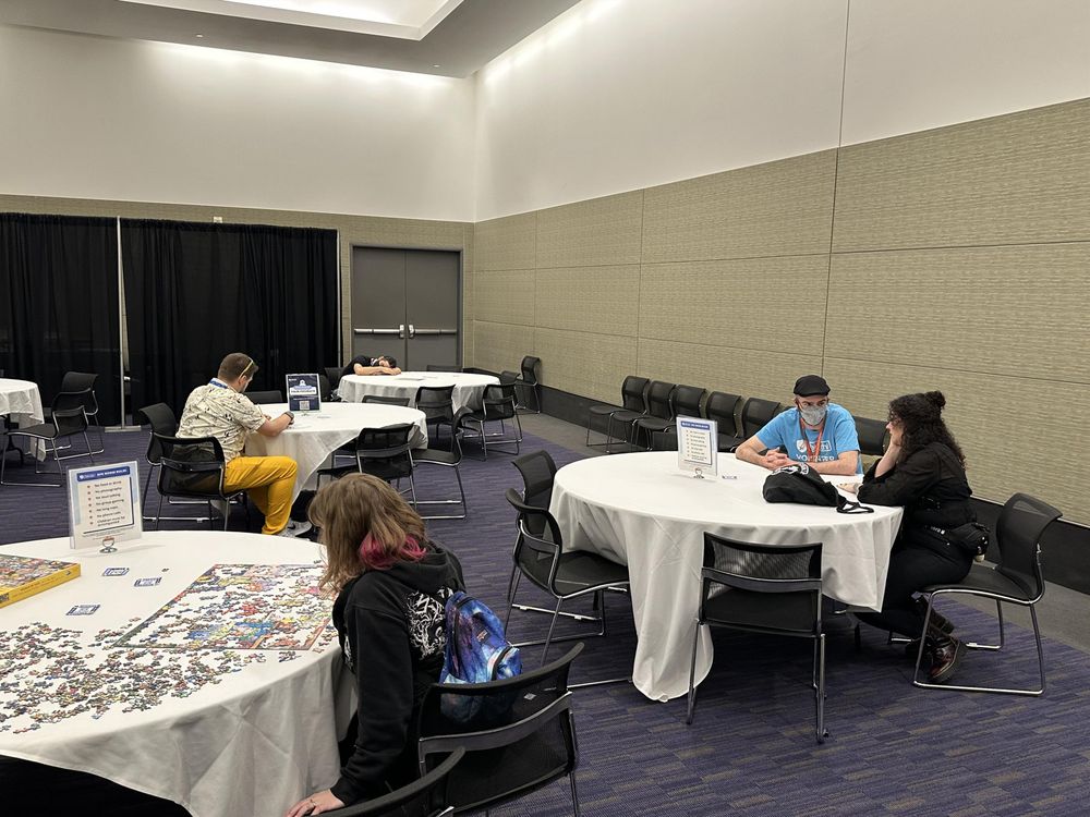 Candid view of an AFK Room. Convention attendees and Take This volunteers sit together at various round tables with white tablecloths. Activities like a jigsaw puzzle are visible on some tables. 