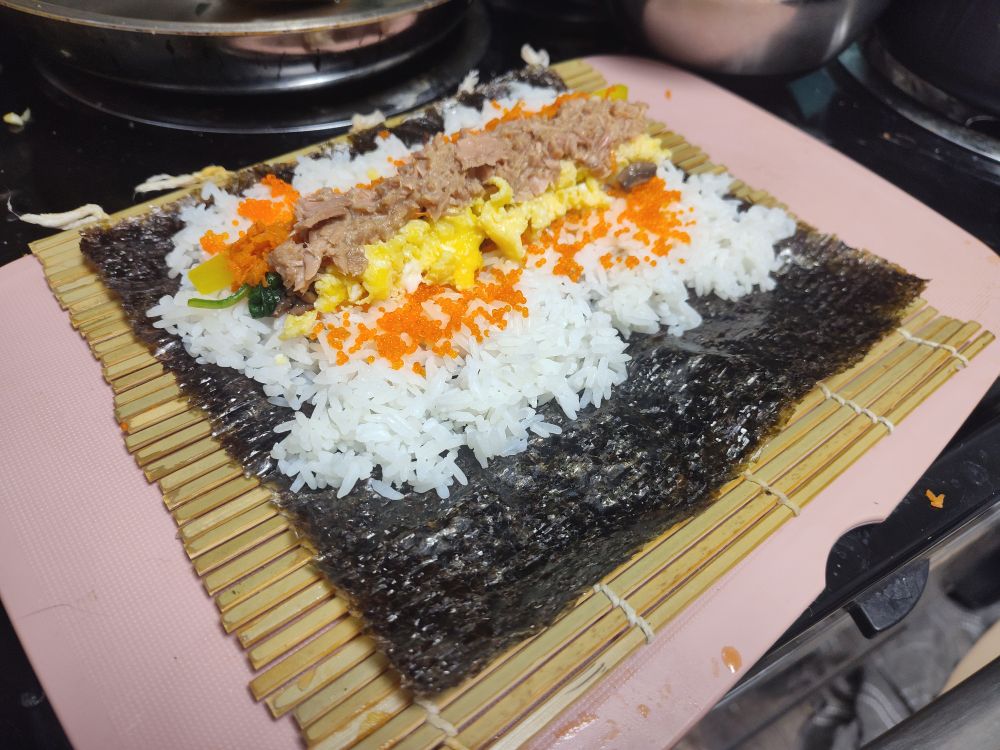 A sheet of seaweed with a pile of rice, tobiko, egg, mushroom, shredded carrot, spinach, takuan and shredded tuna. It is on top of a bamboo sushi roller, itself on top of a cutting board on a stove.