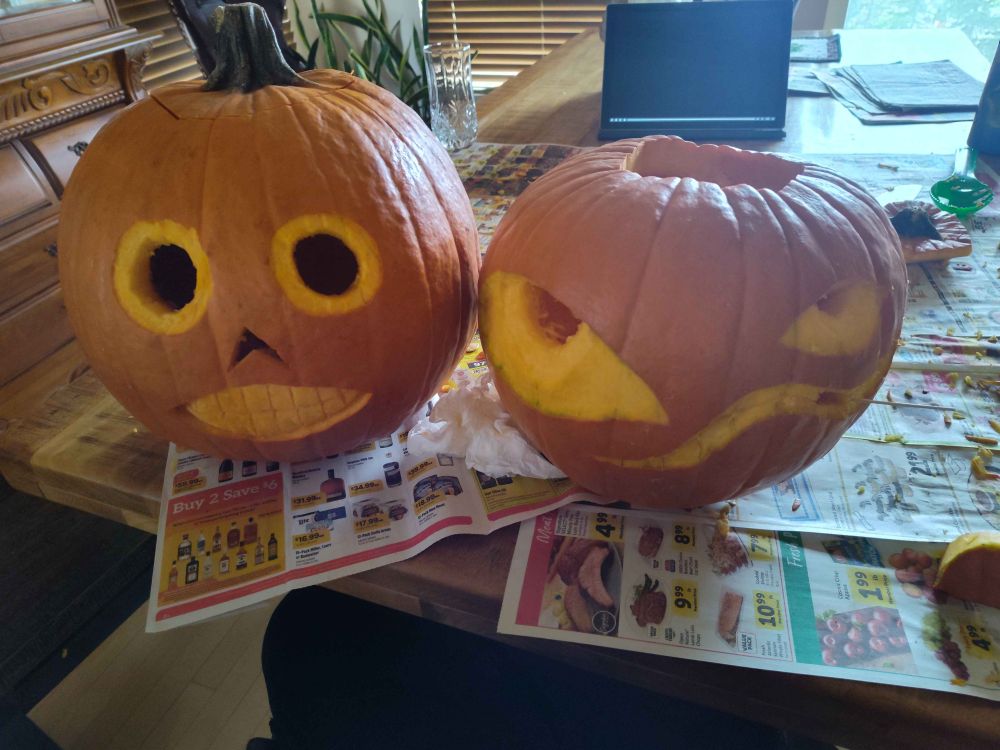Two pumpkins sitting on newspapers on a wooden table. The one on the left is carved with round eyes, a pointy nose, and a big toothy grin resembling Enoch from Over the Garden Wall. The one on the right has two deliberately missized eyes and a twisty grin for a crazed appearance.