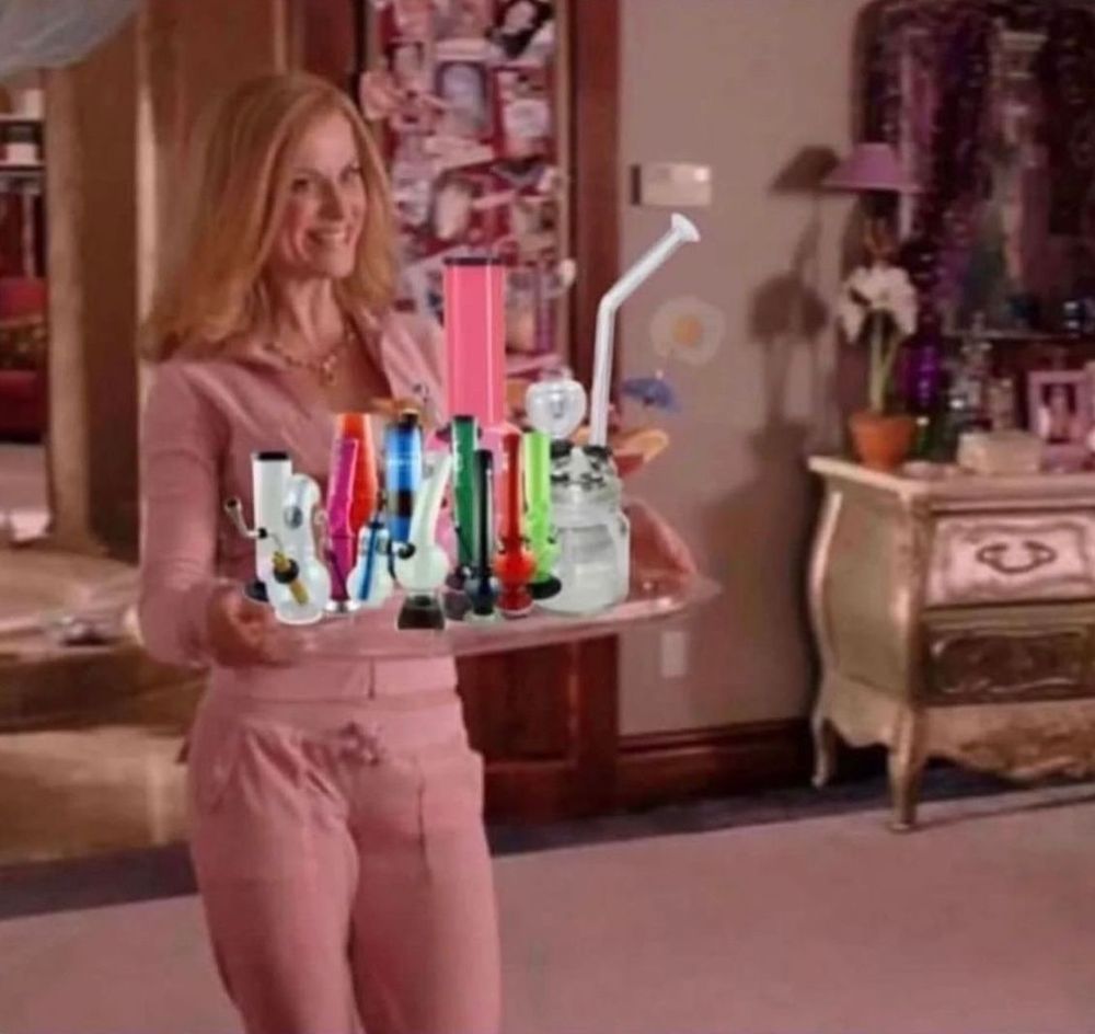 A woman in a pink tracksuit holds a large tray filled with colourful smoking apparatuses, including bongs and pipes, in a well-decorated, cosy room.