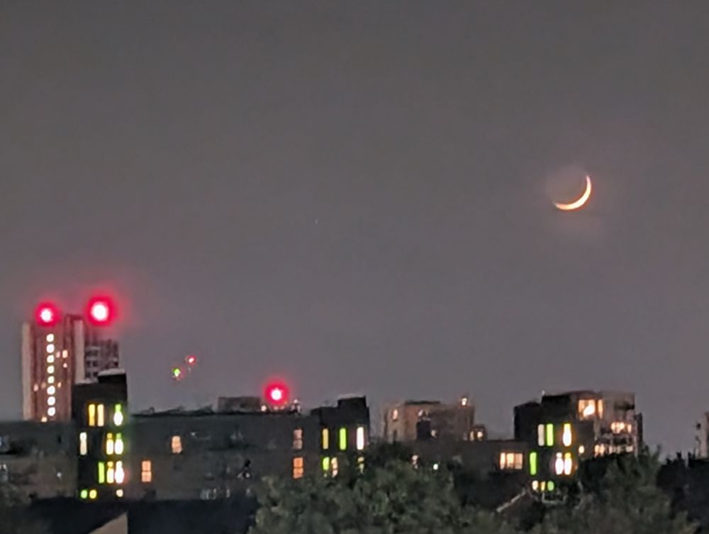 Dark grey night sky. The tops of buildings take up the bottom of the frame. In the sky is the thinnest sliver of a golden moon. 