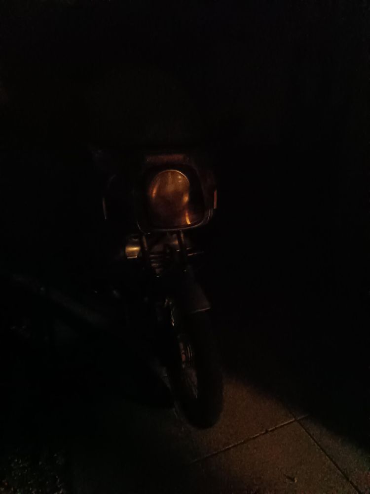 Parked motorbike in the dark, frontwheel and head light visible.