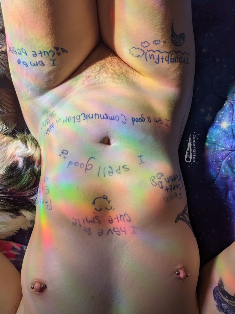 This image features Kit laying on their back fully naked, the sun shining through their window and casting pretty rainbows colors along their tummy. Their pretty pierced titties and natural bush are visible and they are covered in praise focused body writing
