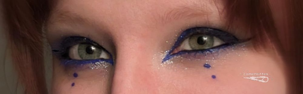 This pic features a cropped photo of Kit's pretty hazel green eyes. Their face is framed by red bangs, very light eye brows, and their make up look consists of metallic blue eyeliner giving them a fun cat eye shape with two blue circles beneath each eye. They have blue glitter eye shadow on their lids, blue mascara, and black liner along their waterline to make their eyes pop.