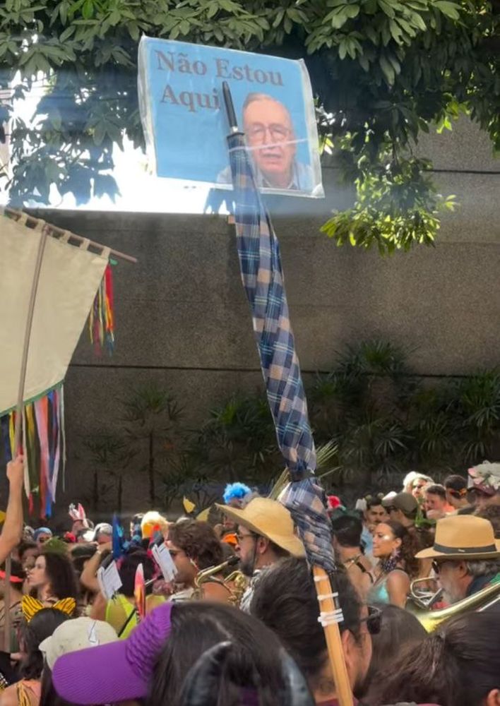 Sombrinha fechada erguida sobre multidao, na.ponta um card amarrado com foto do Olavo de Carvalho e escrito "Não Estou Aqui"