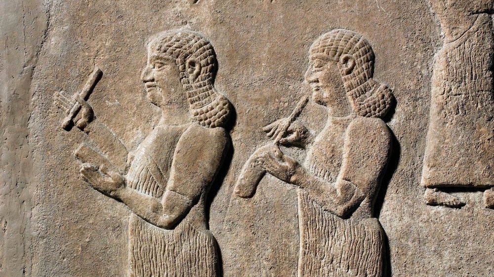 Two scribes depicted in an ancient Assyrian low relief, using styluses to write on clay tablets.