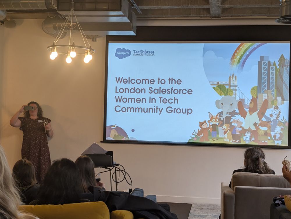 A woman stands in front of a screen holding a phone and drink, appearing to take a photo. The screen displays a slide that reads 'Welcome to the London Salesforce Women in Tech Community Group' with Salesforce mascots and a city skyline illustration. Audience members are seated in the foreground.