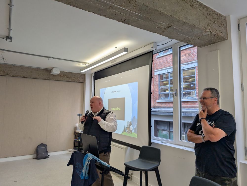 Two speakers presenting at an indoor event, with one speaking into a microphone in front of a laptop and projection screen displaying the GoCardless logo, while the other stands smiling with arms crossed beside a window. 